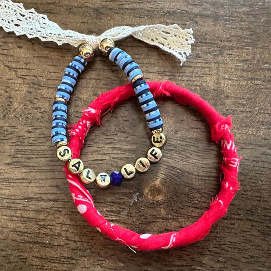 Two handmade beaded bracelets on a dark wood background. One bracelet is bright red with a small white bead accent, and the other is blue with gold letter beads spelling 'LIFE' and blue spacer beads.