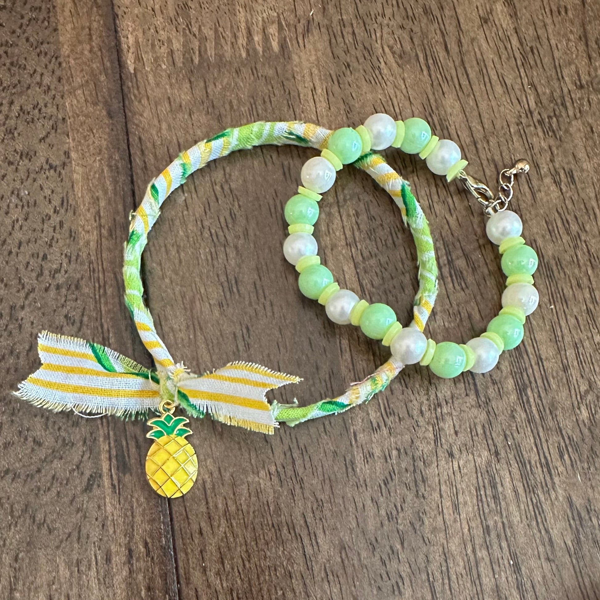 A pair of handmade beaded bracelets with green and white patterned fabric, featuring a yellow pineapple charm and a striped ribbon bow, displayed on a wooden surface.