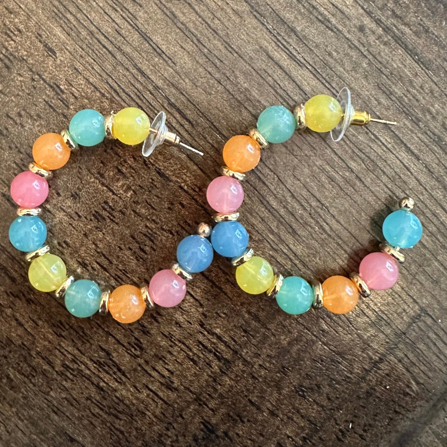 A pair of gold hoop earrings decorated with colorful round beads in pastel shades of pink, orange, yellow, green, and blue, resting on a dark wood surface.