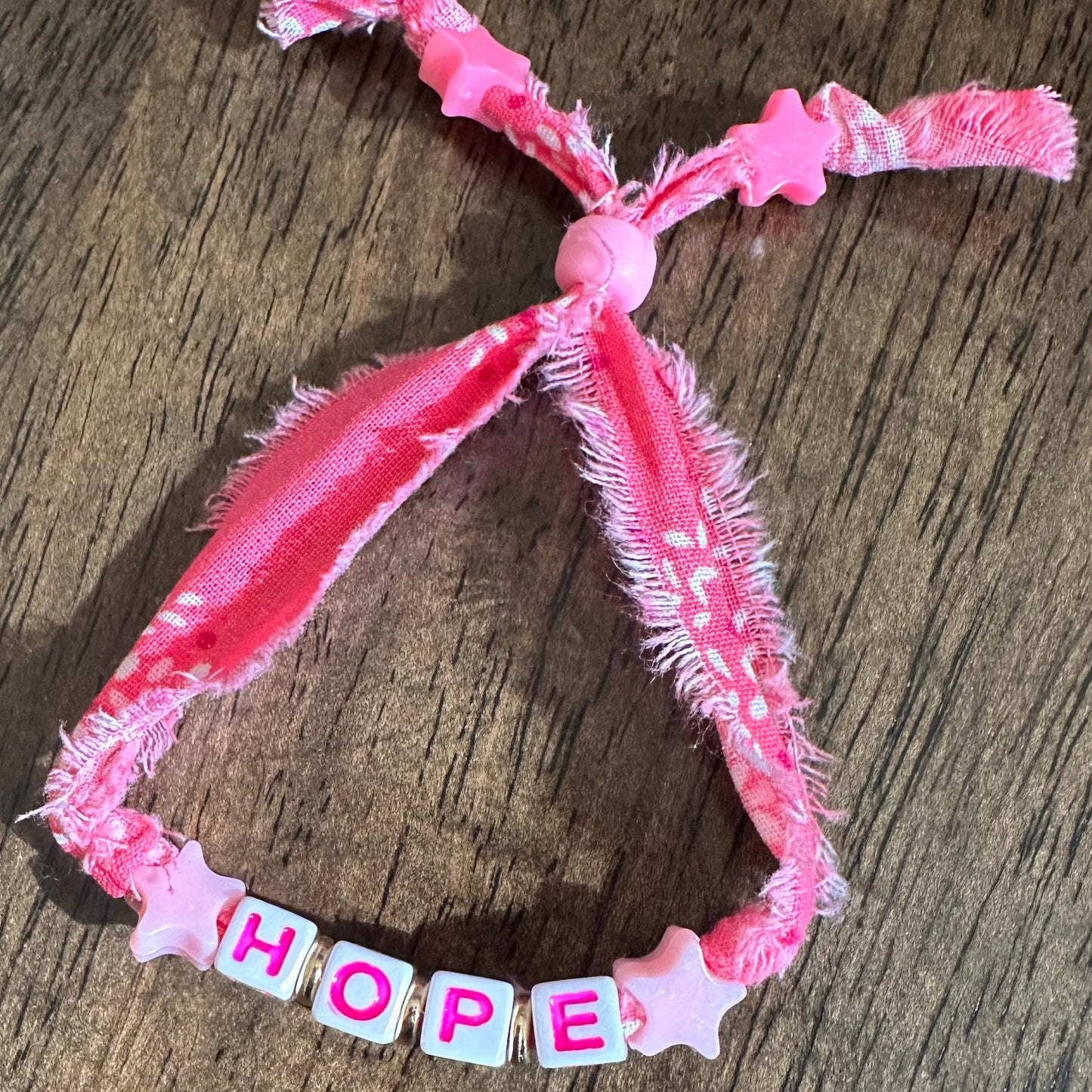A pink fabric pet collar with a knotted top and a beaded chain spelling 'HOPE' on a wooden surface.