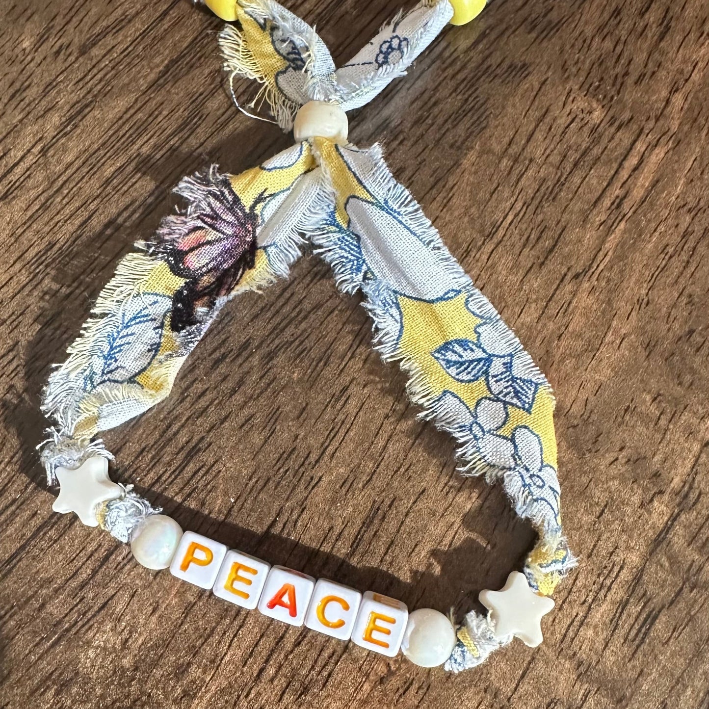 A handmade peace bracelet made of a yellow and white floral fabric, featuring a knotted top with yellow beads, white star charms, and a bottom section spelling 'PEACE' with letter tiles on a wooden surface.