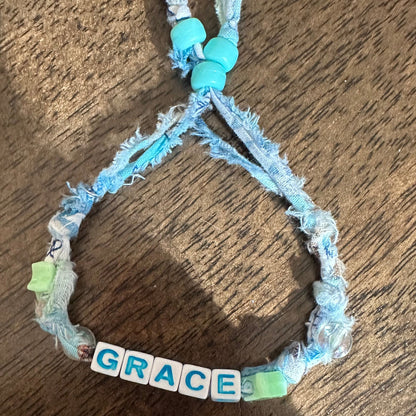 A handmade braided cord bracelet with light blue beads, frayed edges, and letter tiles spelling 'GRACE' on a wooden surface.