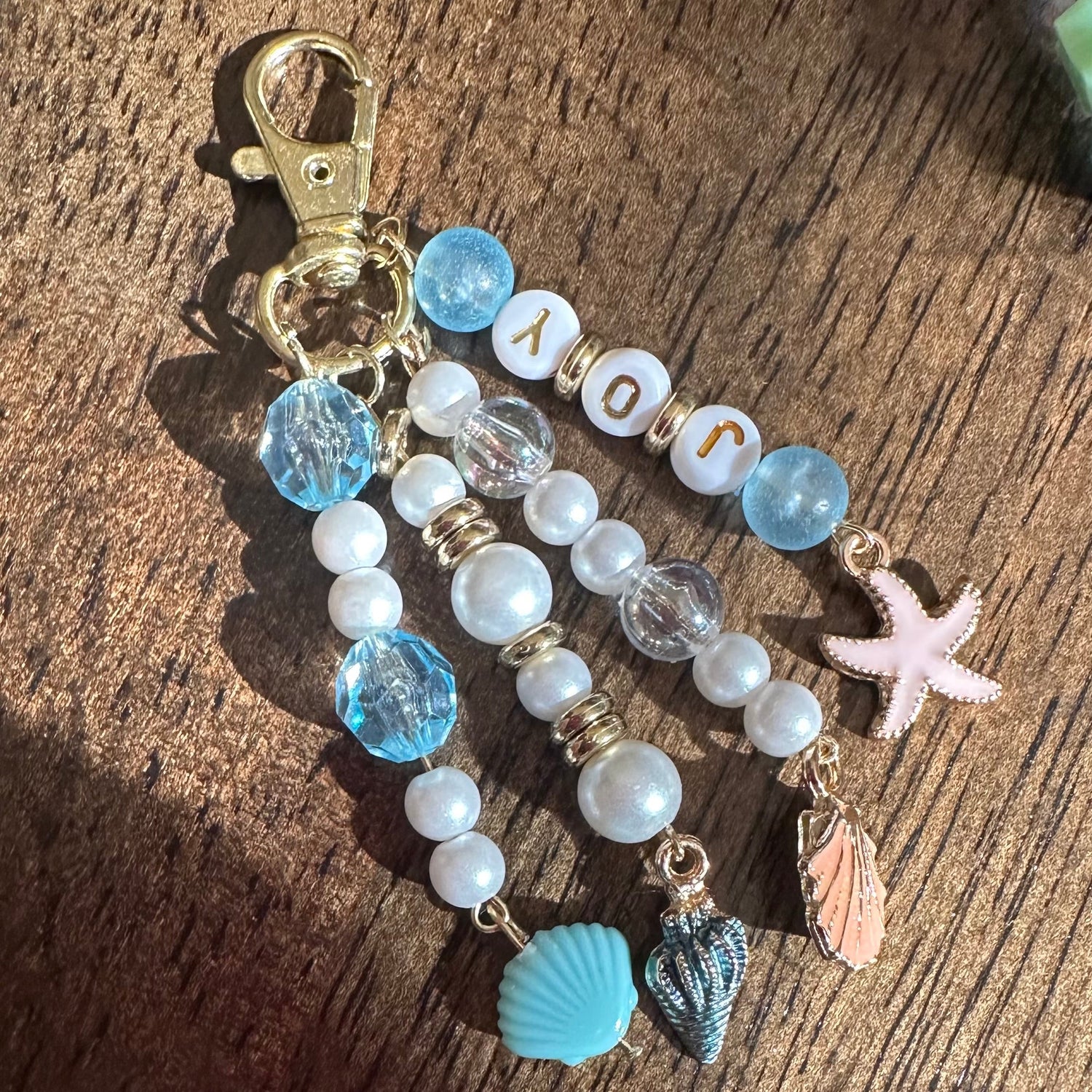 A handmade beaded keychain with a gold clasp, featuring light blue and white beads, letter beads spelling 'JOY', a pink starfish charm, and small shell charms, displayed on a wooden surface.