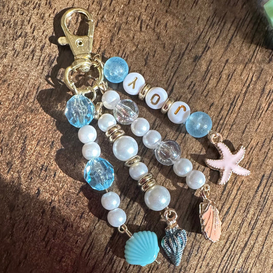 A handmade beaded keychain with a gold clasp, featuring light blue and white beads, letter beads spelling 'JOY', a pink starfish charm, and small shell charms, displayed on a wooden surface.