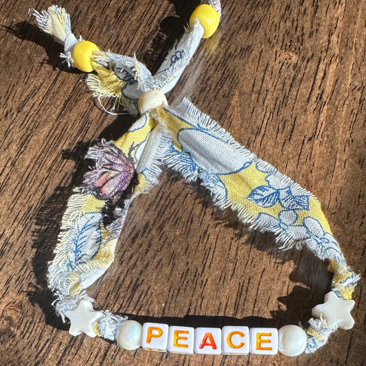 A handmade fabric bracelet with a floral print in yellow, blue, and white, featuring a central row of wooden beads spelling out 'PEACE' in dark orange, yellow, green, and white. The bracelet is displayed on a rustic wooden surface.