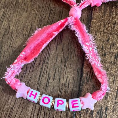 A handmade pink fabric bracelet with frayed edges, featuring four square letter beads spelling 'HOPE' in pink with white outlines, flanked by two pink star beads, displayed on a dark wood background.