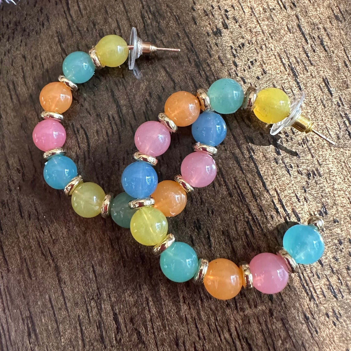 A pair of gold-tone hoop earrings with multicolored round beads in pink, blue, yellow, and orange, displayed on a dark wooden surface.