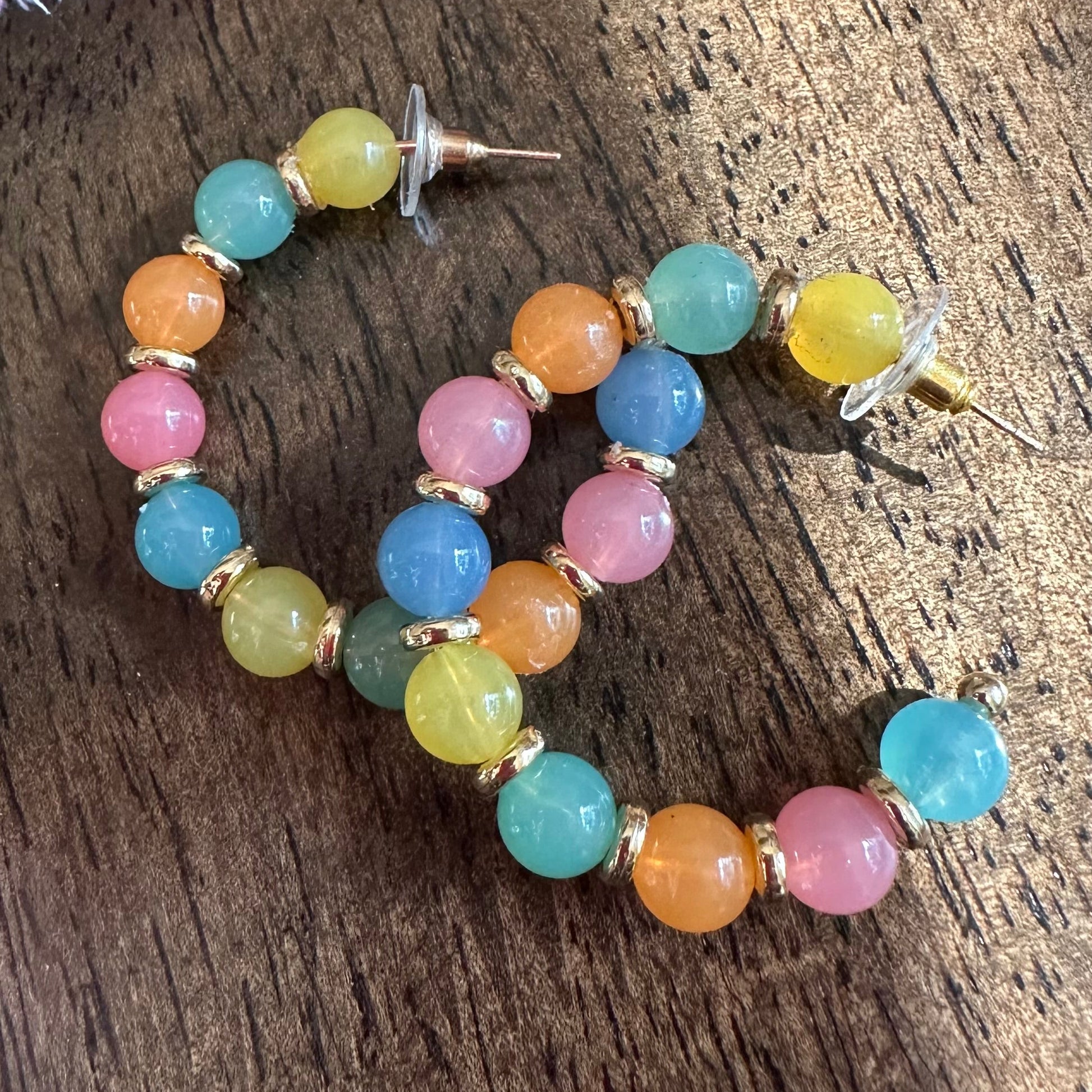 A pair of gold-tone hoop earrings with multicolored round beads in pink, blue, yellow, and orange, displayed on a dark wooden surface.