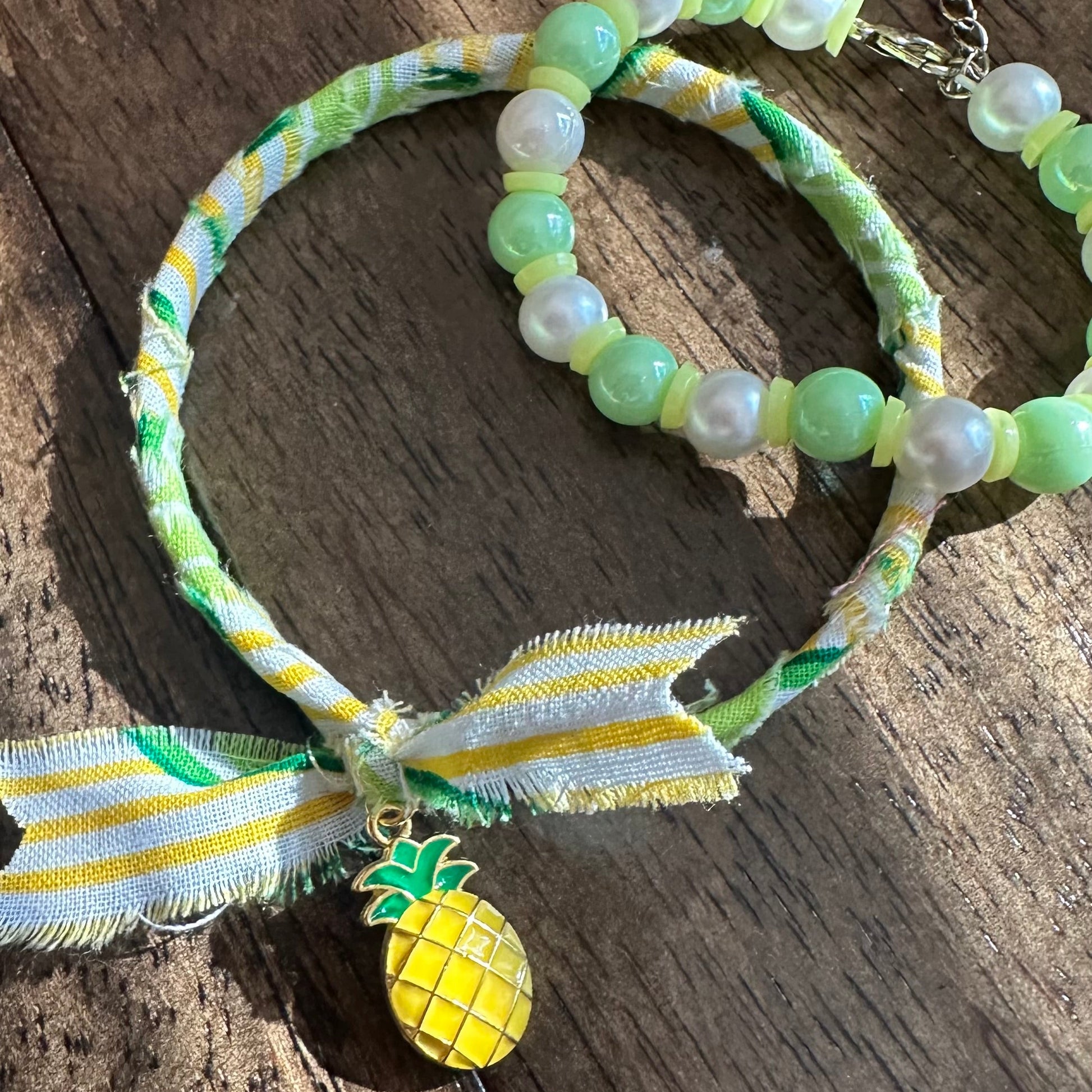 A handmade beaded necklace and matching bracelet featuring light green and white pearls, a green and white patterned fabric wrap, a yellow and white striped ribbon bow, and a yellow pineapple charm, displayed on a rustic wooden surface.