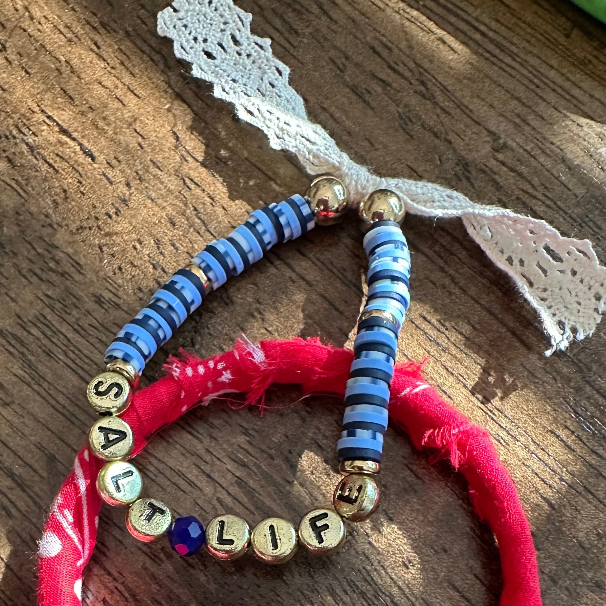 Two handmade fabric and bead bracelets displayed on a rustic wooden surface. One bracelet is made of bright pink frayed fabric with a white lace ribbon and gold letter beads spelling 'LIFEFASHION'.