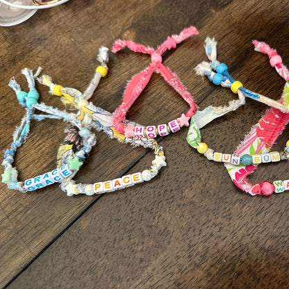 Three handmade fabric and bead bracelets laid out on a dark wooden table. The bracelets feature colorful patterned ribbons, small beads, and letter beads spelling out words like HOPE, SUN BUM, PEACE, and. GRACE.