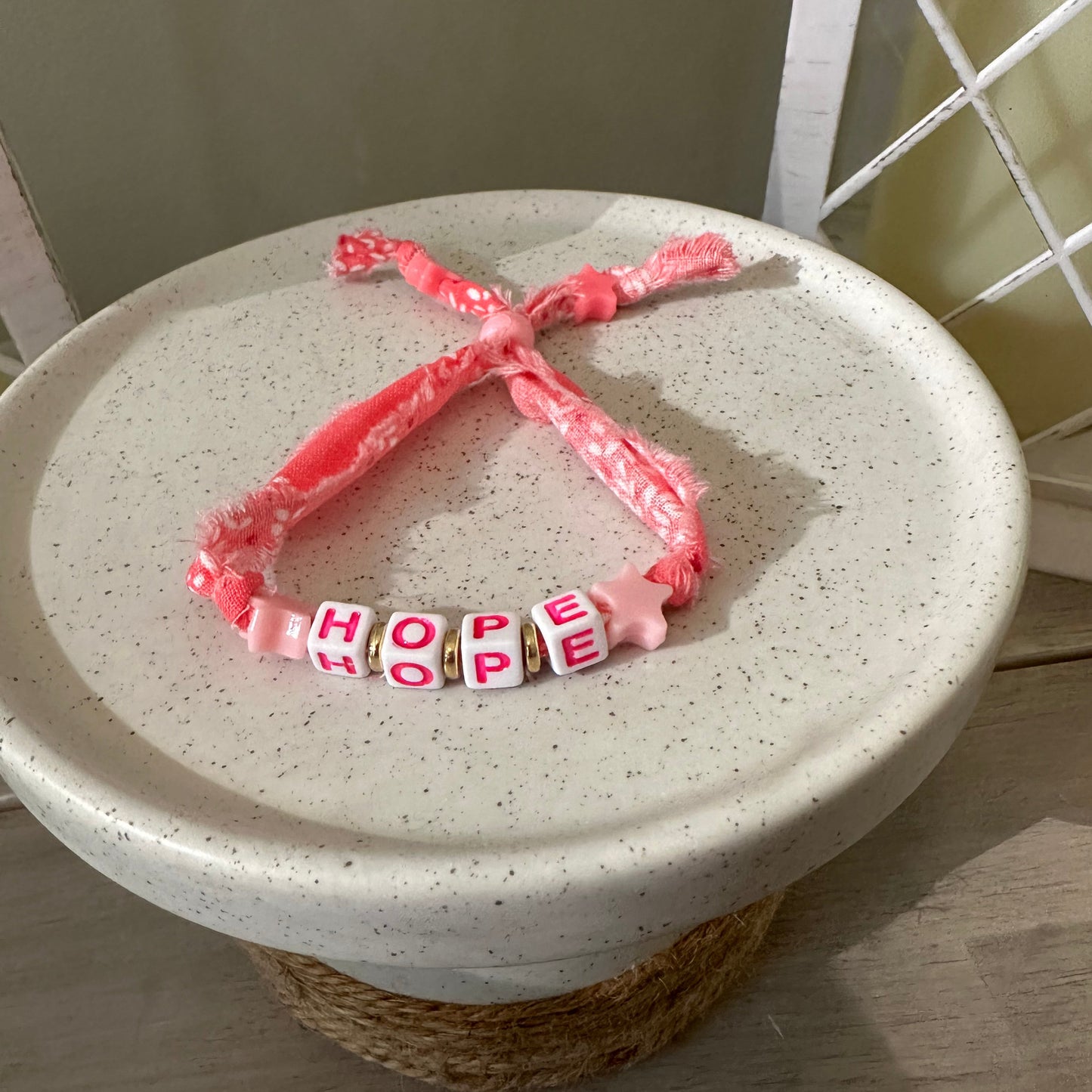 A pink beaded bracelet with a knotted ribbon end, displayed on a speckled white ceramic plate. The beads spell out 'HOPE' in white with pink lettering.