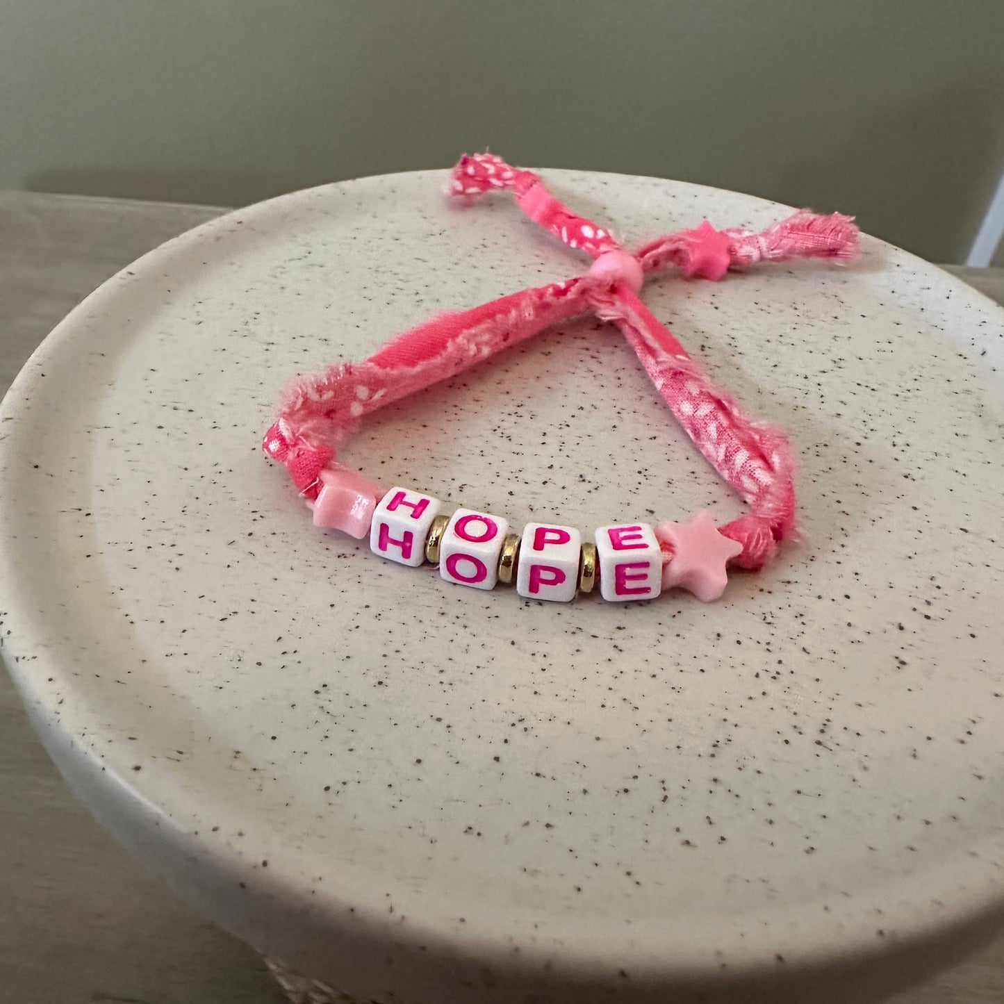 A pink braided cord bracelet with four letter beads spelling 'HOPE'  in pink letters on white beads, resting on a speckled cream ceramic plate.