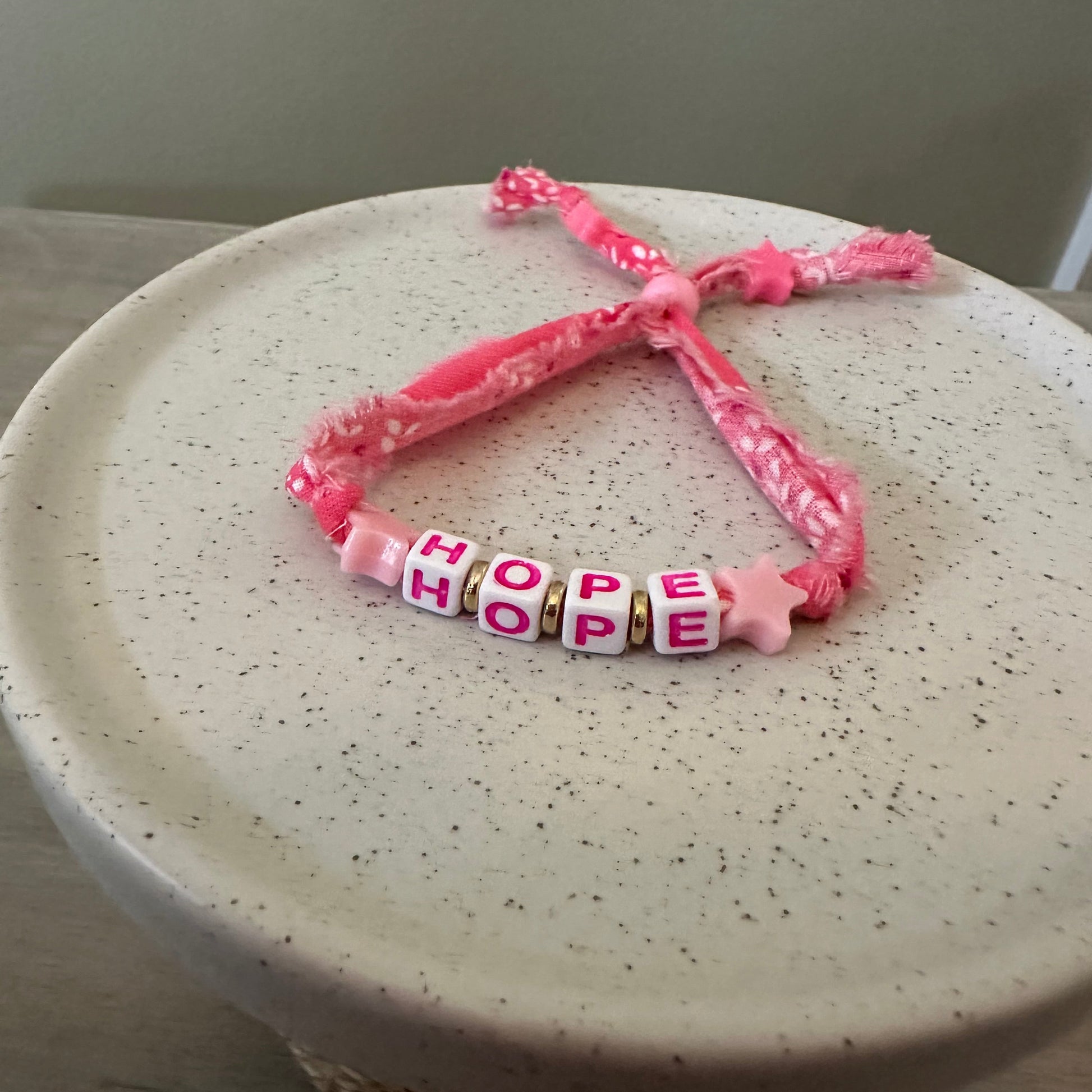 A pink braided cord bracelet with four letter beads spelling 'HOPE'  in pink letters on white beads, resting on a speckled cream ceramic plate.