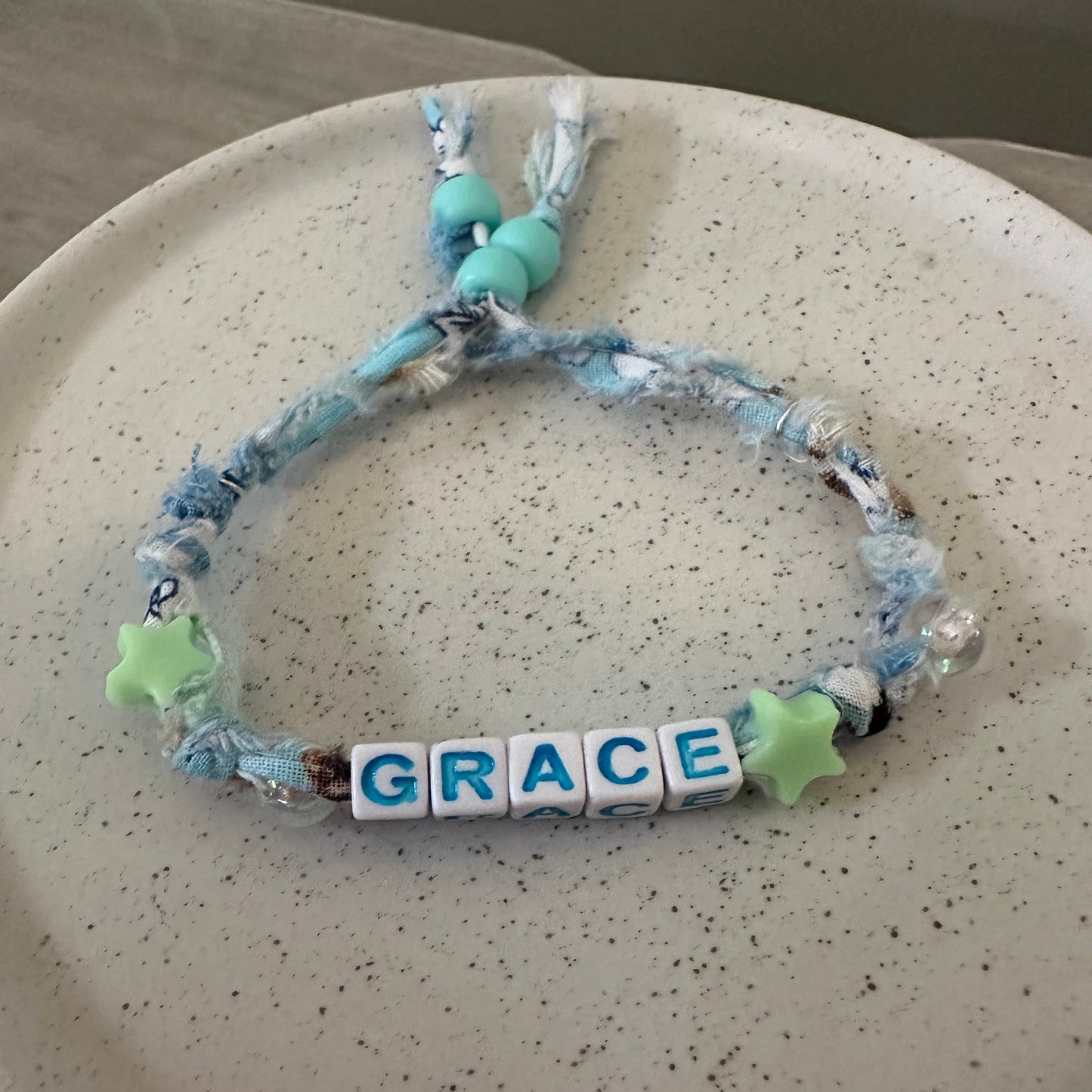 A handmade beaded bracelet with blue and white marbled cord, light blue beads, green star charms, and white letter tiles spelling 'GRACE', displayed on a speckled beige ceramic plate.