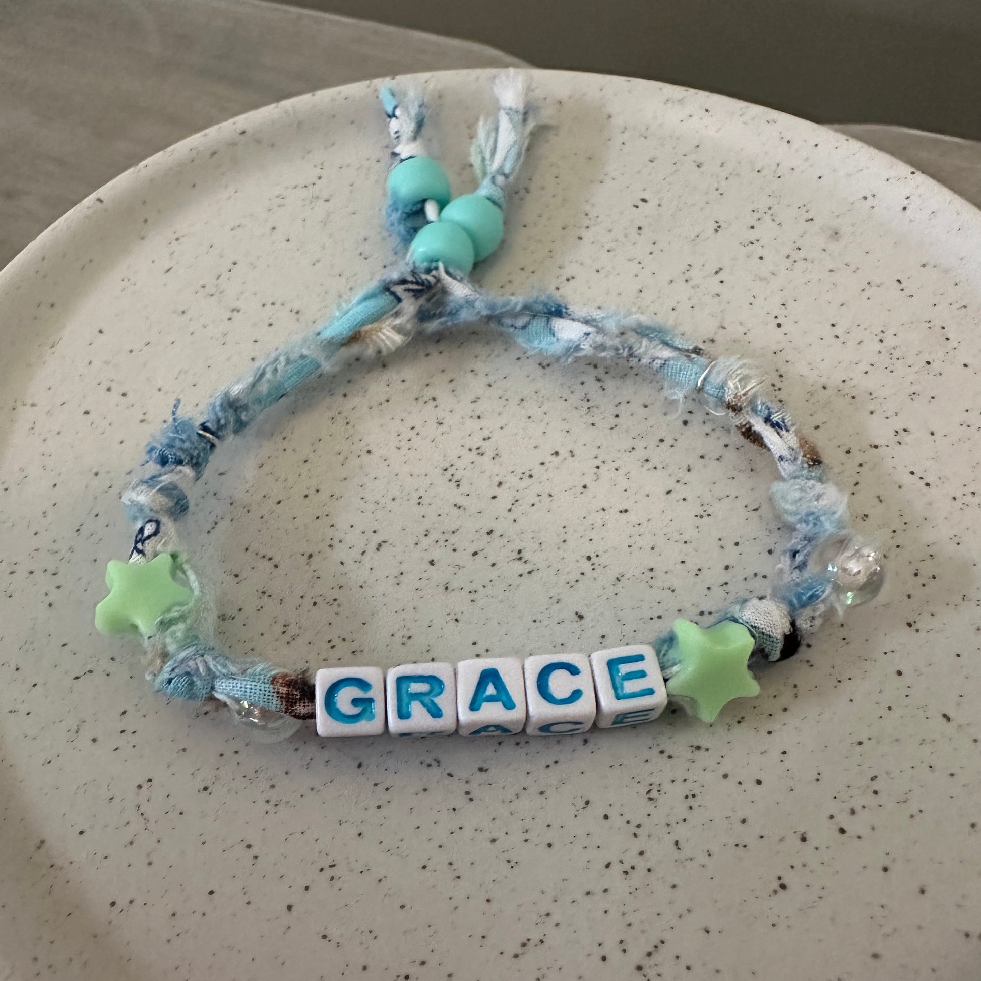 A handmade beaded bracelet with blue and white marbled cord, light blue beads, green star charms, and white letter tiles spelling 'GRACE', displayed on a speckled beige ceramic plate.