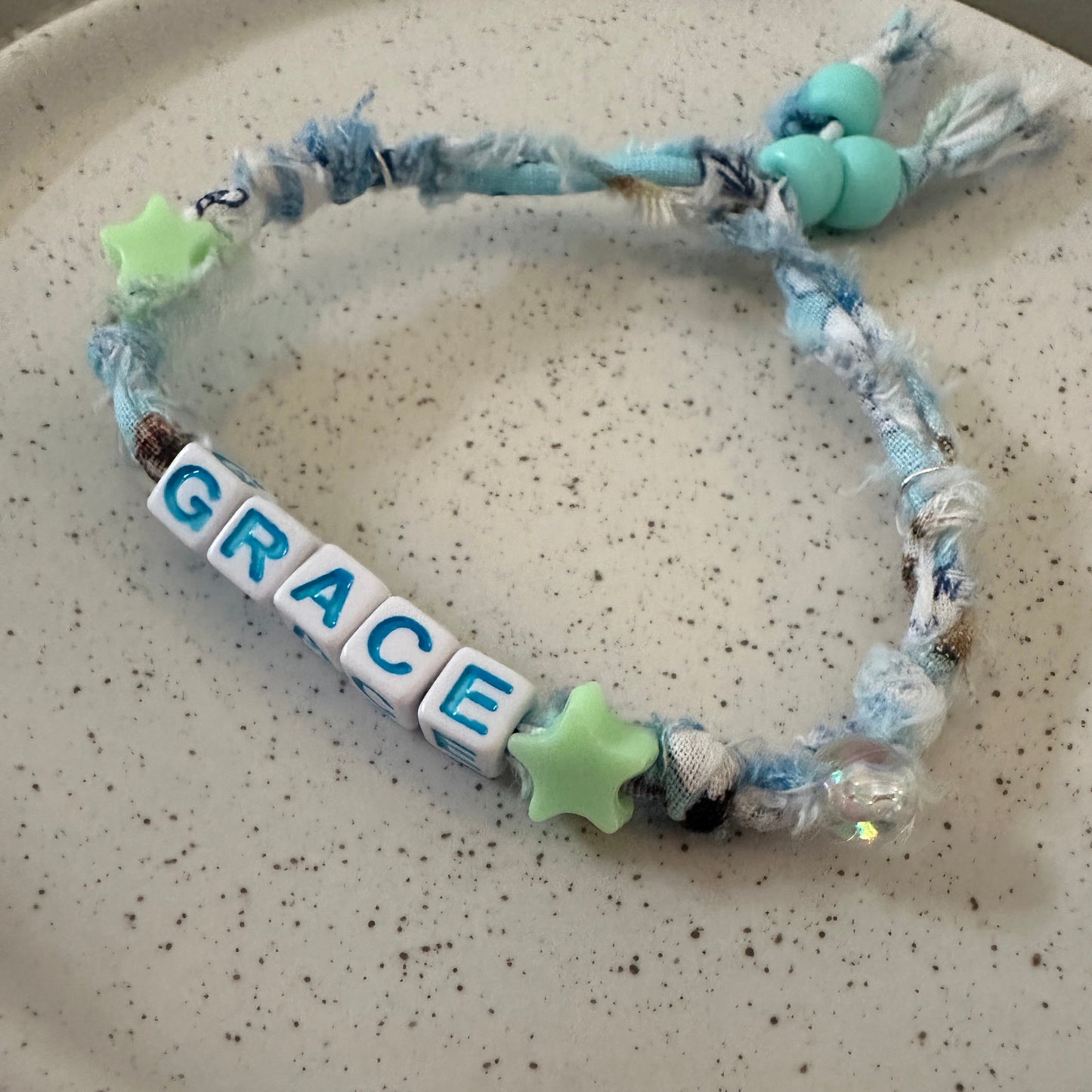 A handmade fabric bracelet with a tassel, blue and white yarn, light green star beads, and white letter tiles spelling 'GRACE', displayed on a speckled beige ceramic plate.