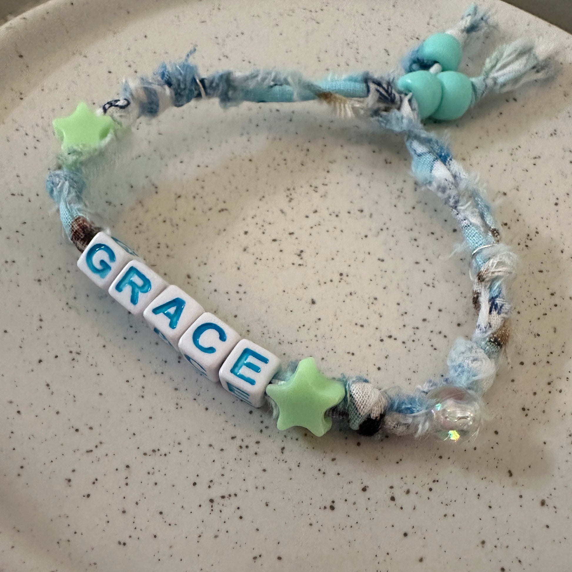 A handmade fabric bracelet with a tassel, blue and white yarn, light green star beads, and white letter tiles spelling 'GRACE', displayed on a speckled beige ceramic plate.
