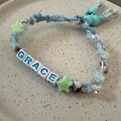 A handmade fabric bracelet with a tassel, blue and white yarn, light green star beads, and white letter tiles spelling 'GRACE', displayed on a speckled beige ceramic plate.