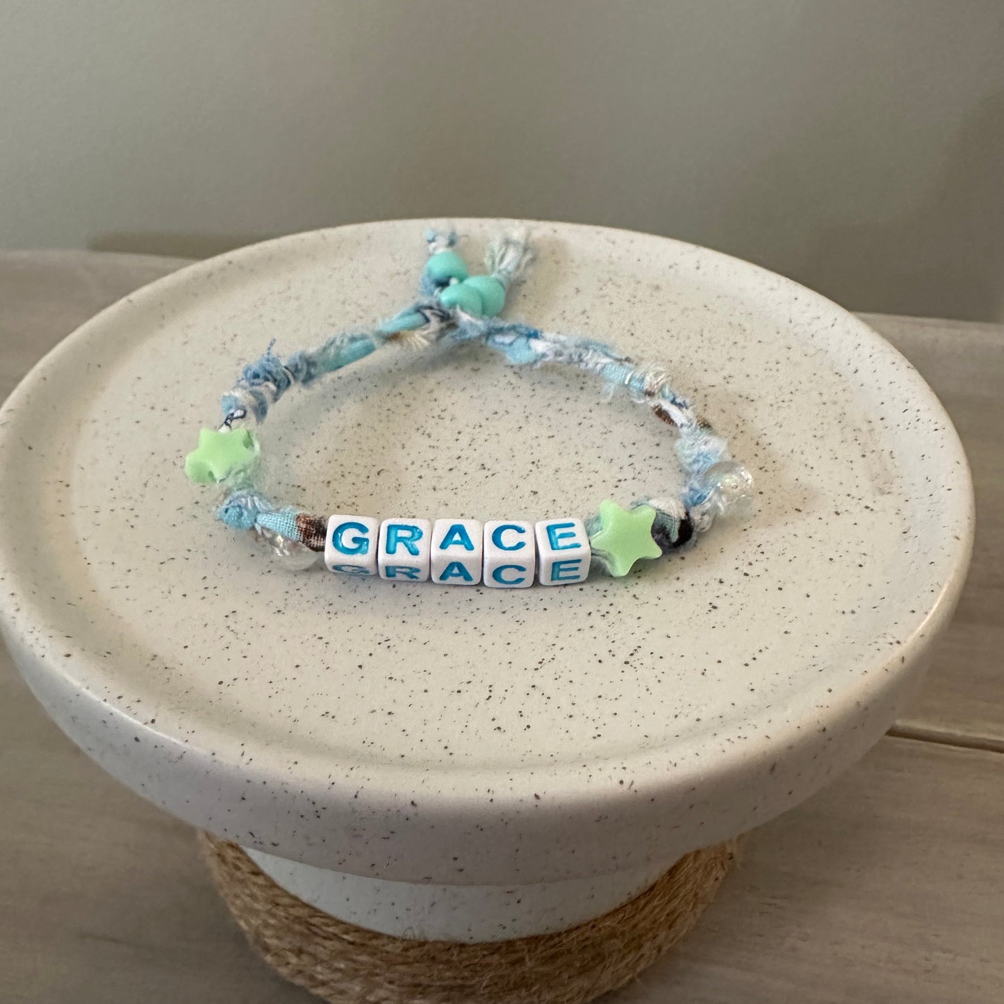 A handmade beaded bracelet with light blue and white marbled beads, a white cube bead spelling 'GRACE', and two mint green star beads, displayed on a speckled ceramic stand.