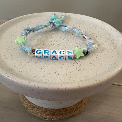 A handmade beaded bracelet with blue and white frayed cord, light blue beads, and white cube beads spelling 'GRACE', displayed on a speckled ceramic stand.