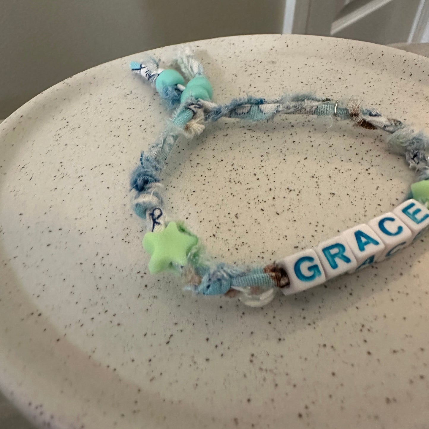 A handmade beaded bracelet with blue and white frayed rope, a white letter bead spelling 'GRACE', and a light green star charm, resting on a speckled beige ceramic plate.