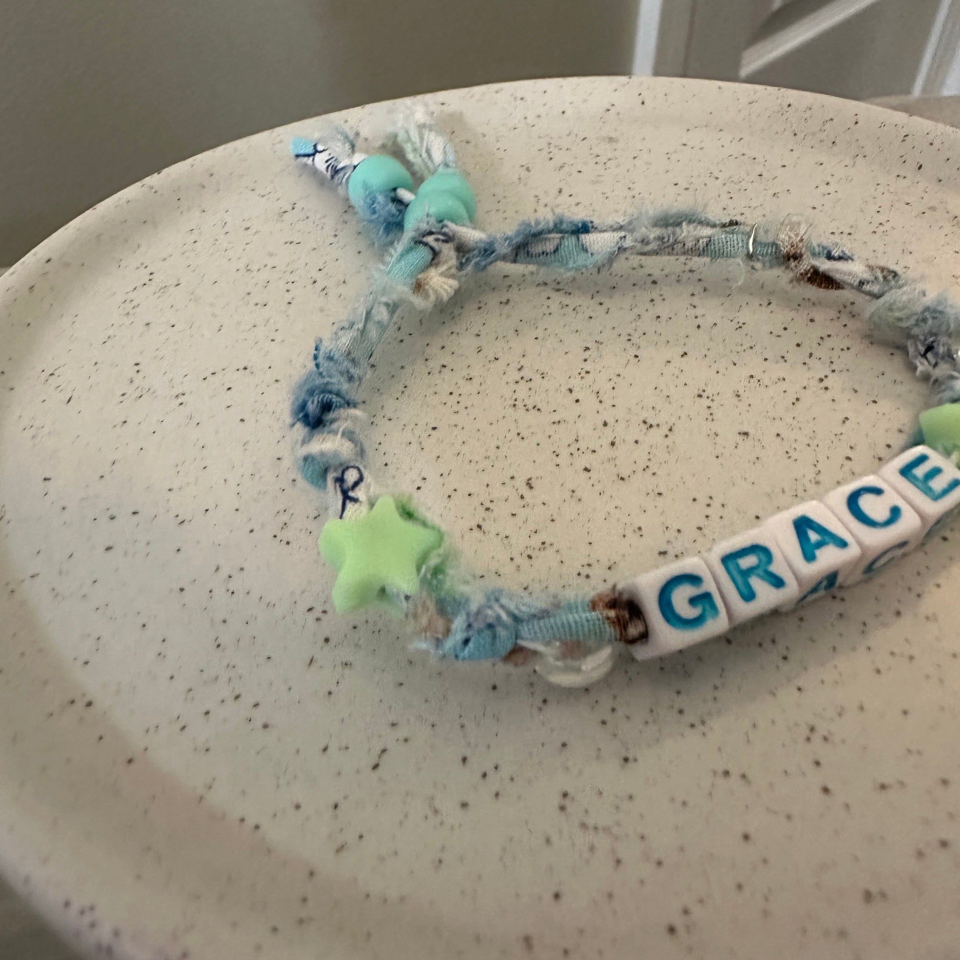 A handmade beaded bracelet with blue and white frayed rope, a white letter bead spelling 'GRACE', and a light green star charm, resting on a speckled beige ceramic plate.
