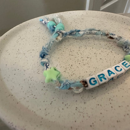 A handmade beaded bracelet with blue and white frayed rope, a white letter bead spelling 'GRACE', and a light green star charm, resting on a speckled beige ceramic plate.