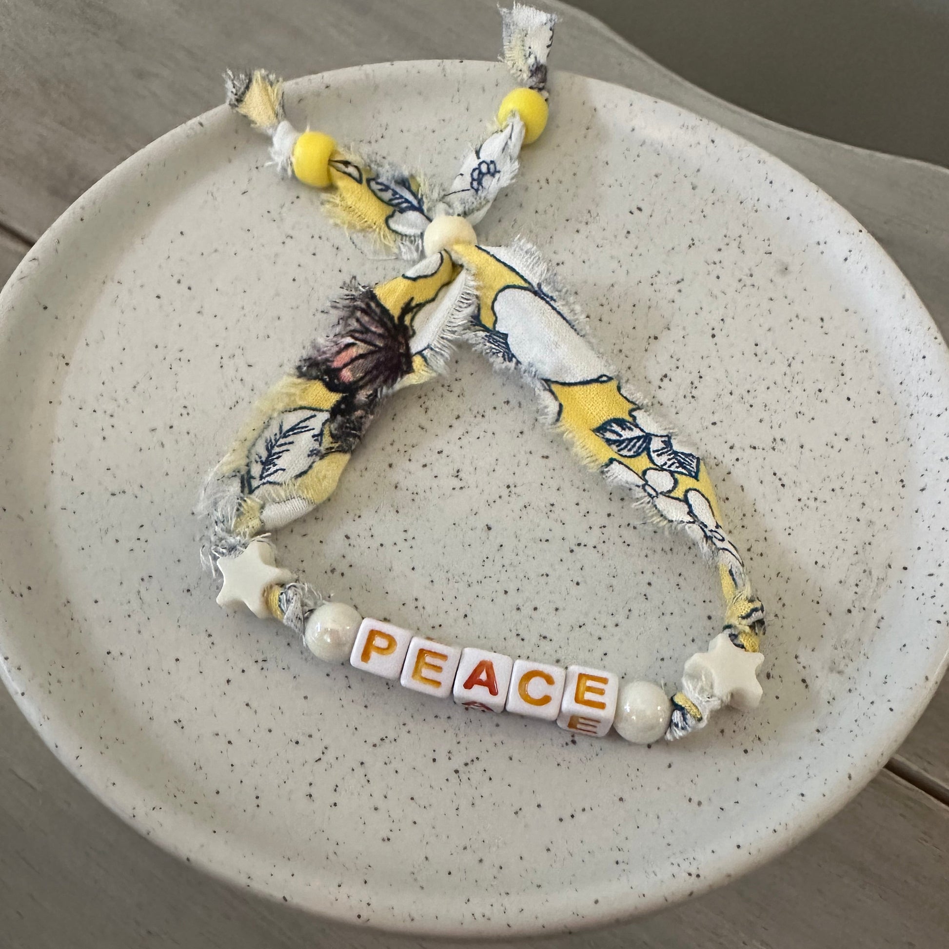 A handmade fabric bracelet with a distressed yellow and white floral print, featuring white star beads spelling out 'PEACE' on a speckled white ceramic plate.