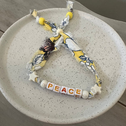 A handmade fabric bracelet with a distressed yellow and white floral print, featuring white star beads spelling out 'PEACE' on a speckled white ceramic plate.