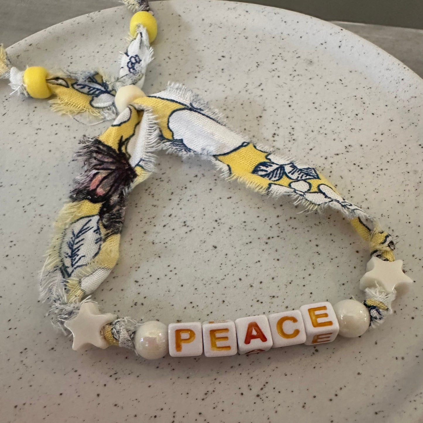 A handmade beaded bracelet with a yellow and white patterned fabric strap, white star beads, and wooden cube beads spelling out 'PEACE', displayed on a speckled ceramic plate.