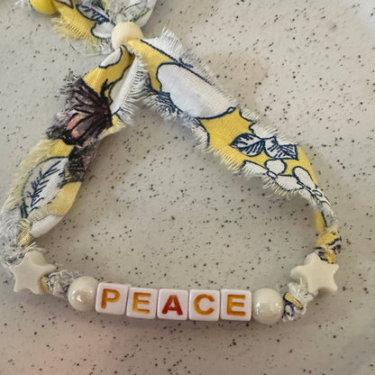 A handmade beaded bracelet featuring a yellow, white, and navy floral fabric ribbon, white star beads, and white cube beads spelling out the word 'PEACE'. The bracelet is displayed on a speckled beige ceramic plate.