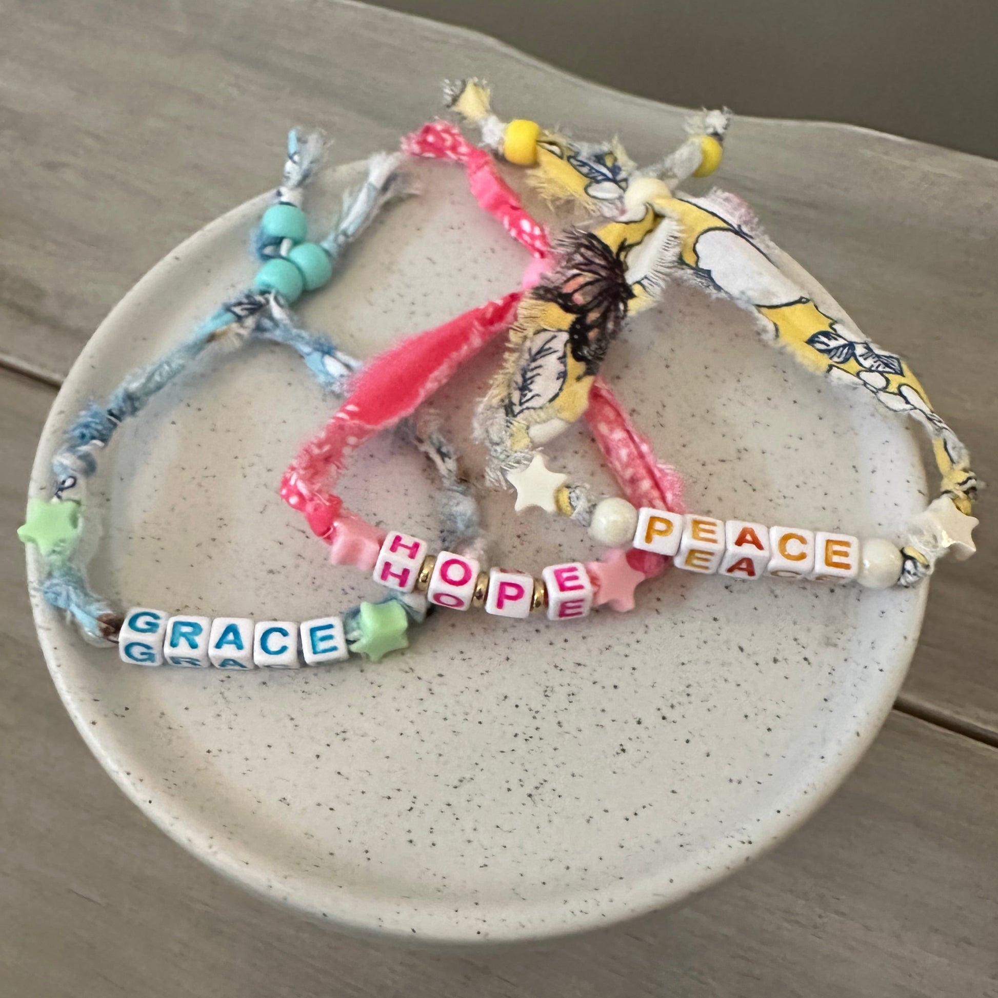 A collection of handmade beaded and fabric bracelets arranged on a speckled ceramic plate, placed on a light wood surface. The bracelets feature colorful beads, letter tiles spelling out words like 'PEACE', 'HOPE', and 'GRACE', along with pink ribbon, yellow patterned paper, and blue denim accents.