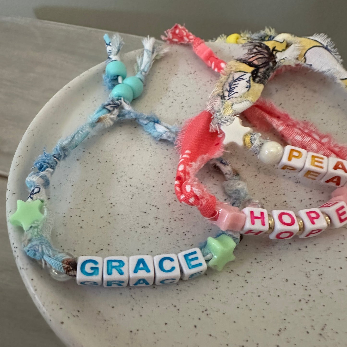 A close-up of several handmade beaded bracelets arranged on a speckled off-white plate. The bracelets include white letter beads spelling 'PEACE', 'HOPE', and 'GRACE', along with pink patterned fabric beads, light blue beads, green star beads, and multicolored decorative elements.