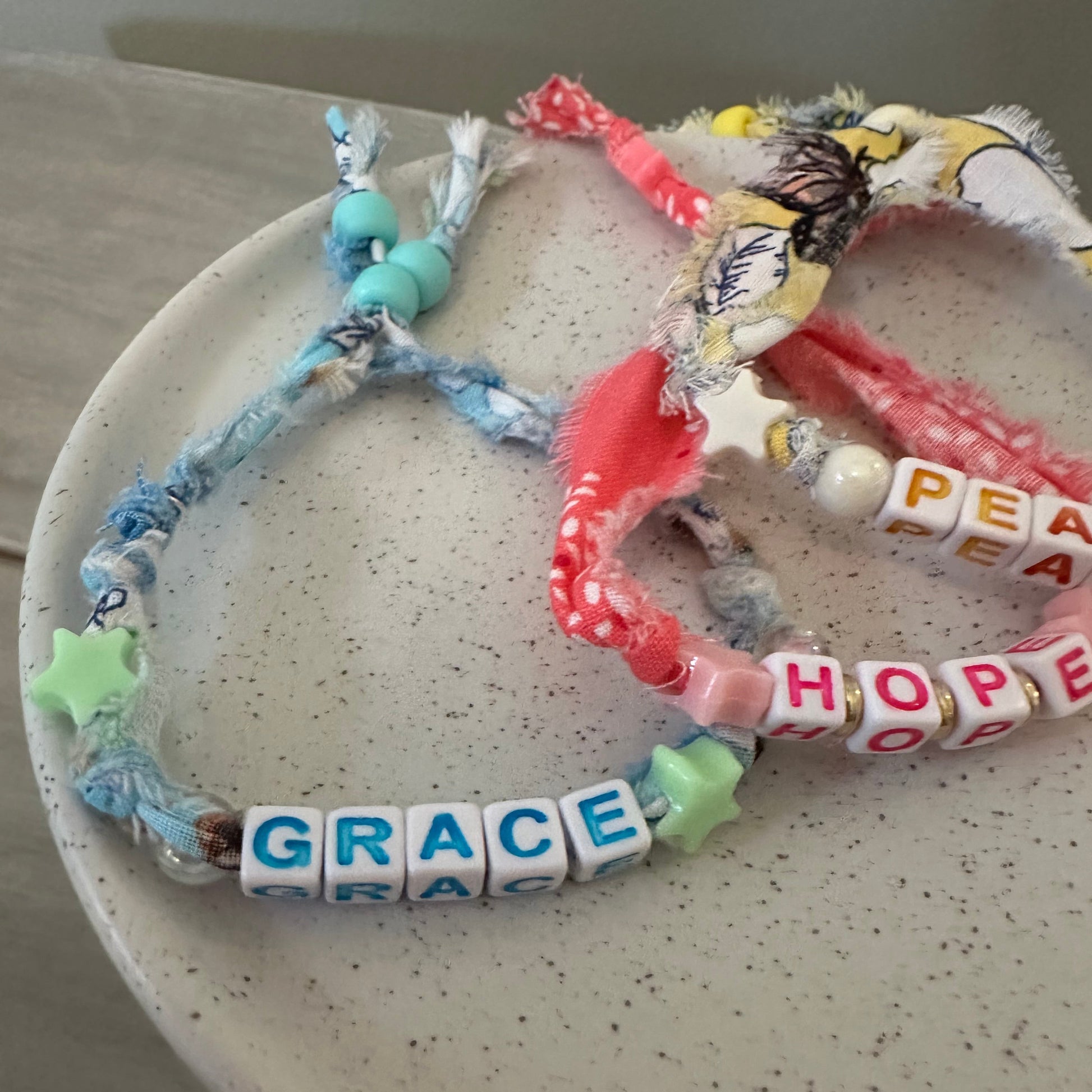 A close-up of several handmade beaded bracelets arranged on a speckled off-white plate. The bracelets include white letter beads spelling 'PEACE', 'HOPE', and 'GRACE', along with pink patterned fabric beads, light blue beads, green star beads, and multicolored decorative elements.