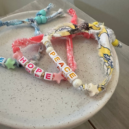 A handmade heart-shaped beaded bracelet made from colorful scrapbook fabrics and letter beads spelling out PEACE and HOPE, displayed on a speckled ceramic dish.