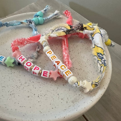 A handmade heart-shaped beaded bracelet made from colorful scrapbook fabrics and letter beads spelling out PEACE and HOPE, displayed on a speckled ceramic dish.