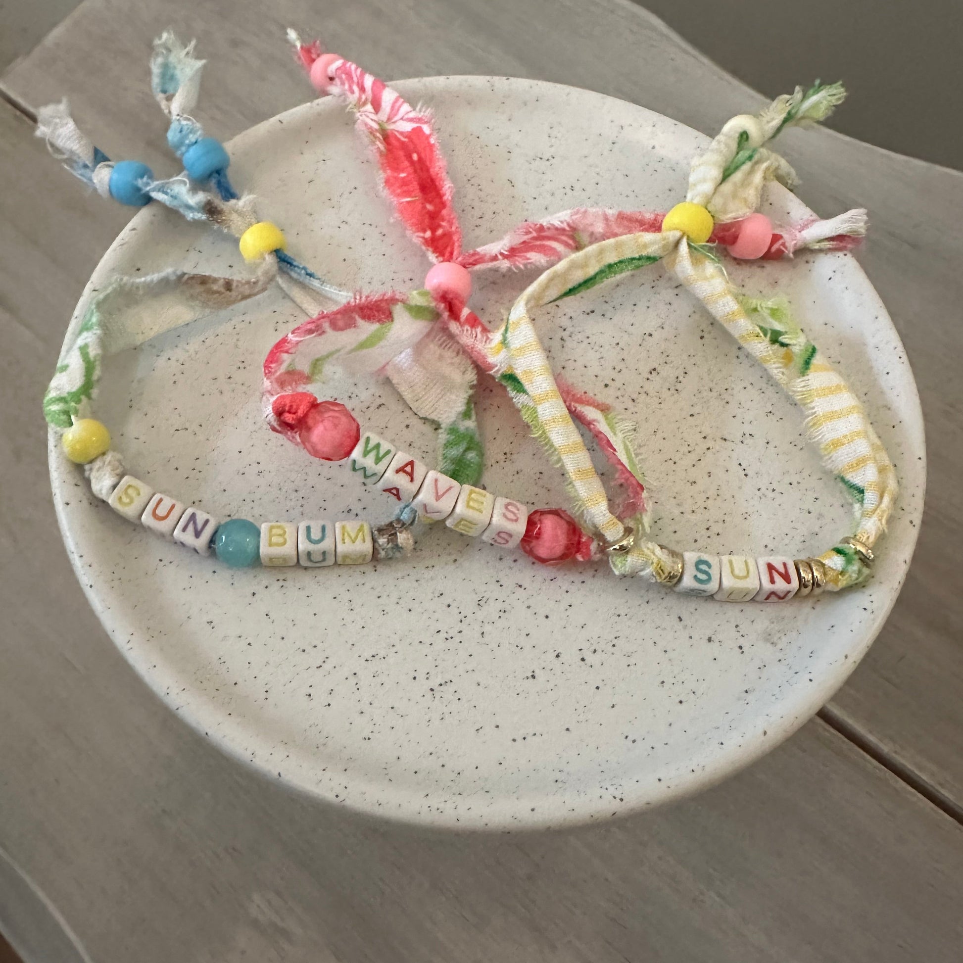 Three handmade fabric beaded bracelets with colorful tassels and letter beads spelling 'BUNN' and 'WAVE', displayed on a speckled ceramic plate over a gray wooden surface.