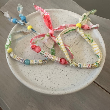 Three handmade fabric beaded bracelets with colorful tassels and letter beads spelling 'BUNN' and 'WAVE', displayed on a speckled ceramic plate over a gray wooden surface.