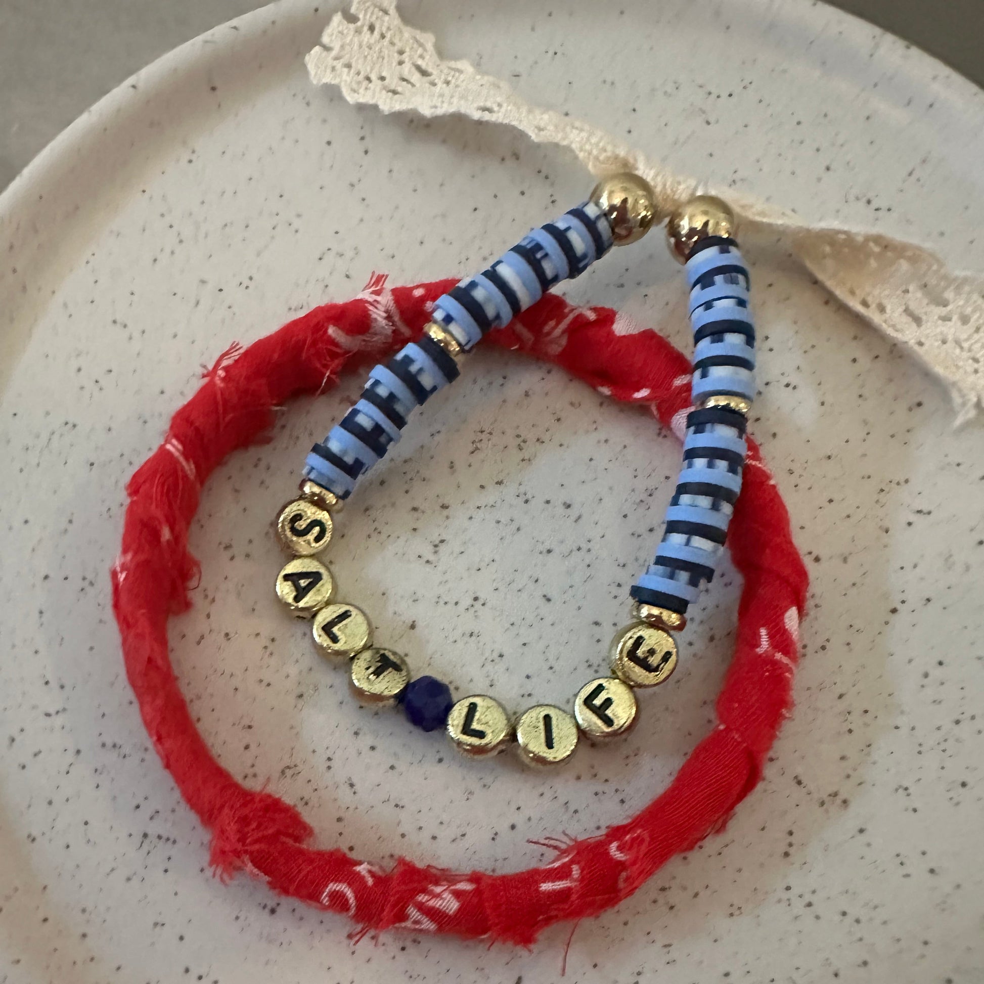 A handmade beaded bracelet set on a speckled white ceramic plate. The set features a red frayed fabric bracelet, a blue and white striped fabric bracelet, gold letter beads spelling  salt life', and blue and gold spacer beads.