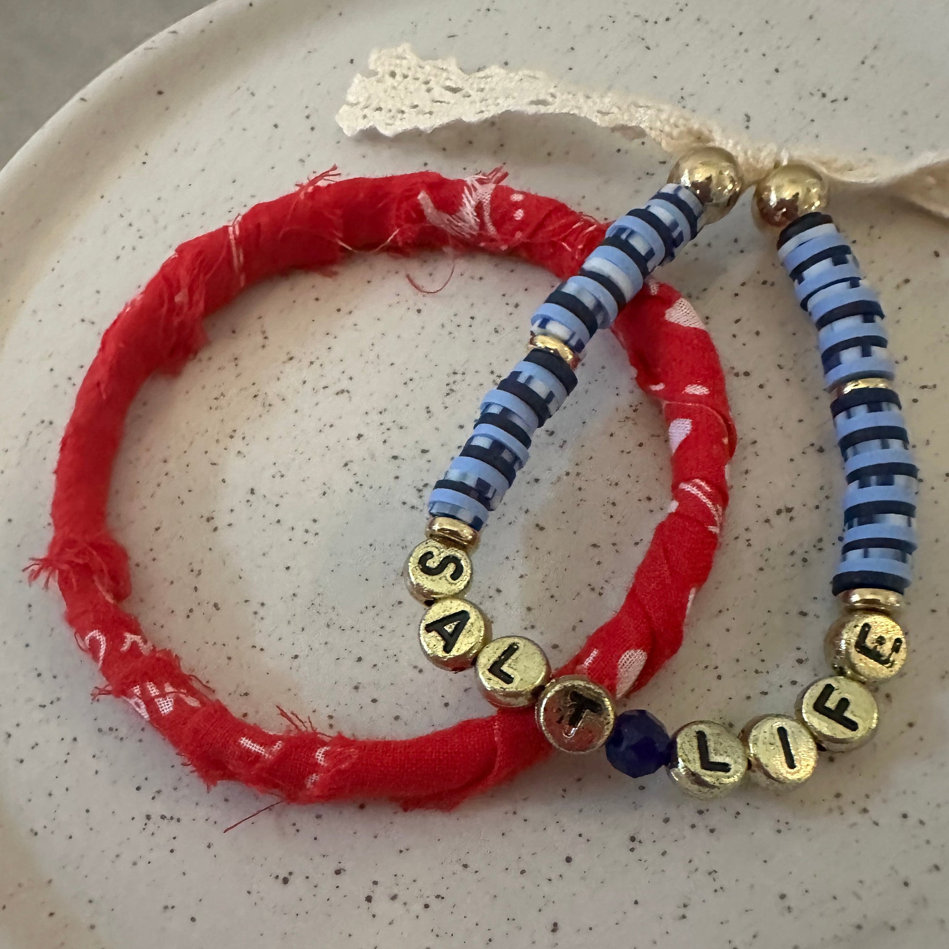 Two handmade beaded bracelets displayed on a speckled white ceramic plate. One bracelet is made of alternating blue and white striped beads with gold letter beads spelling ' SALT LIFE ' (partially obscured by a red bracelet).