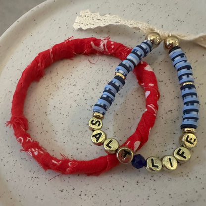 Two handmade beaded bracelets displayed on a speckled white ceramic plate. One bracelet is made of alternating blue and white striped beads with gold letter beads spelling ' SALT LIFE ' (partially obscured by a red bracelet).