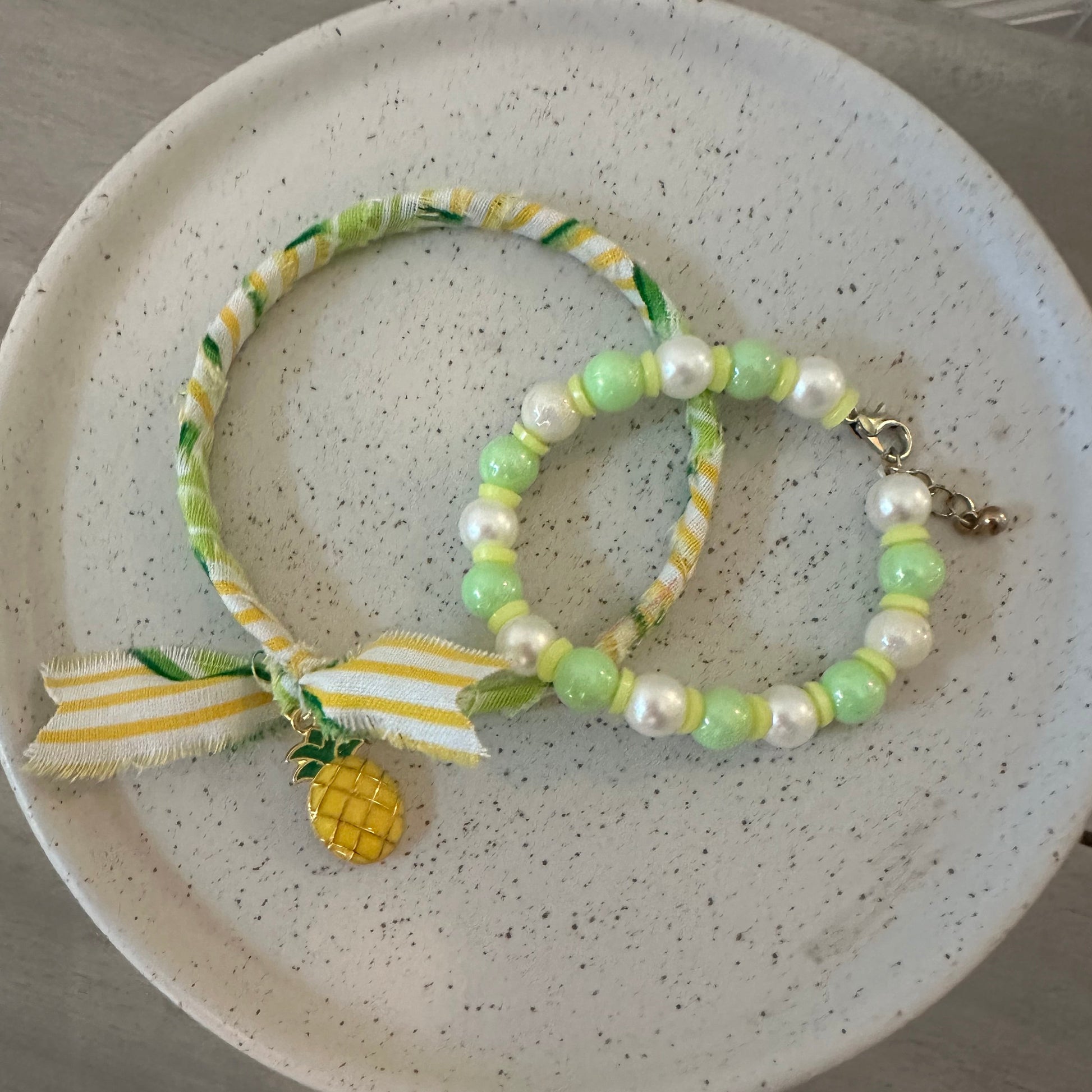 A set of two beaded and ribbon bracelets in pastel green, white, and yellow, displayed on a speckled ceramic plate.