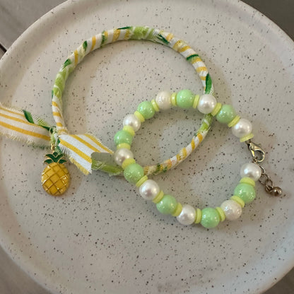 A handmade beaded bracelet and matching wristlet strap featuring light green and white faux pearls, yellow-green patterned fabric, a yellow pineapple charm, and a white ribbon, displayed on a speckled ceramic plate.