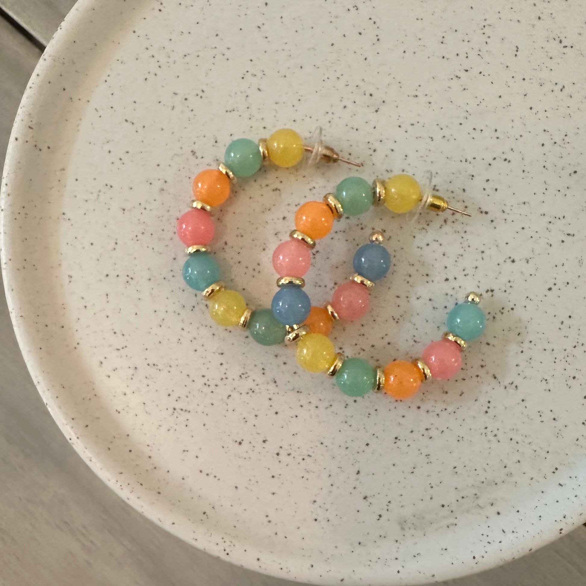 A pair of pastel beaded earrings with gold-toned metal findings, displayed on a speckled white ceramic plate over a light wood surface.