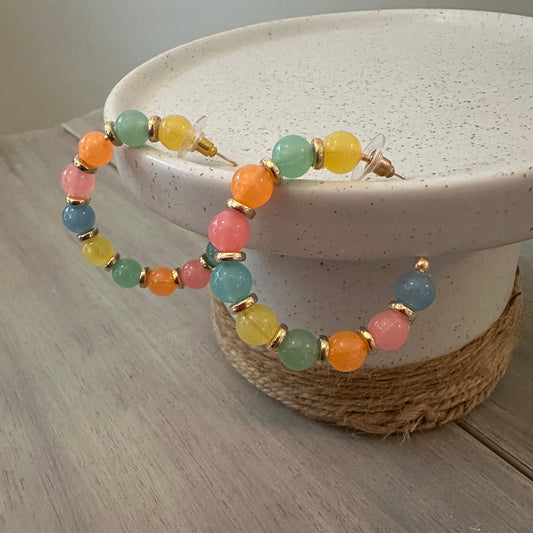 A pair of beaded bracelets with pastel multicolor round beads and gold-tone metal findings, displayed on a white speckled jewelry stand against a light wood background.