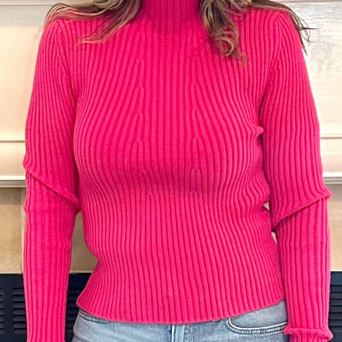 Person wearing a bright pink ribbed sweater and light blue jeans, standing indoors.