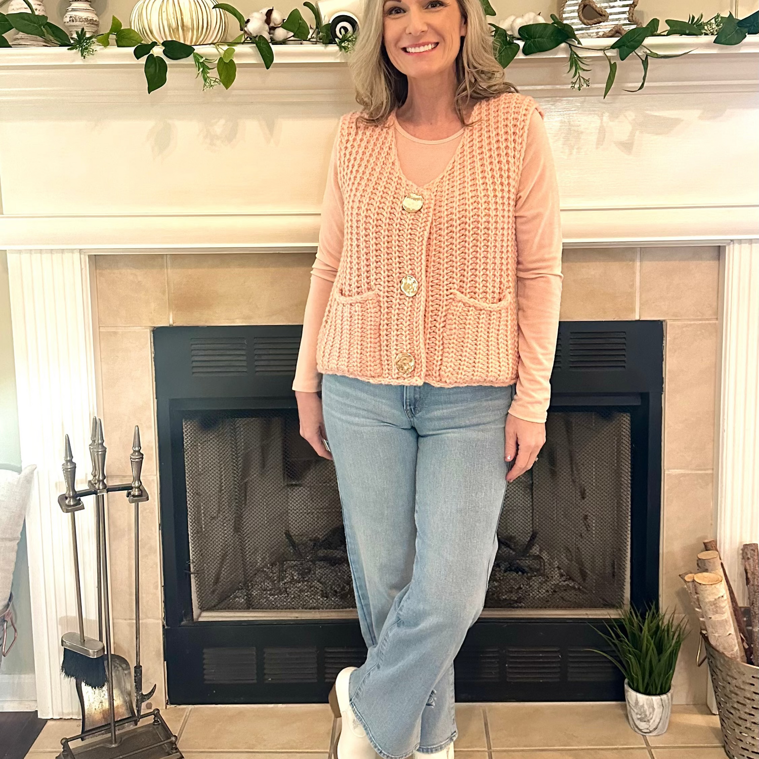 Woman wearing a pink cardigan and blue jeans standing in front of a fireplace.