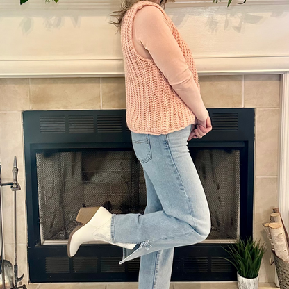 Woman in a pink sweater and jeans standing in front of a fireplace with decorative items on the mantel.