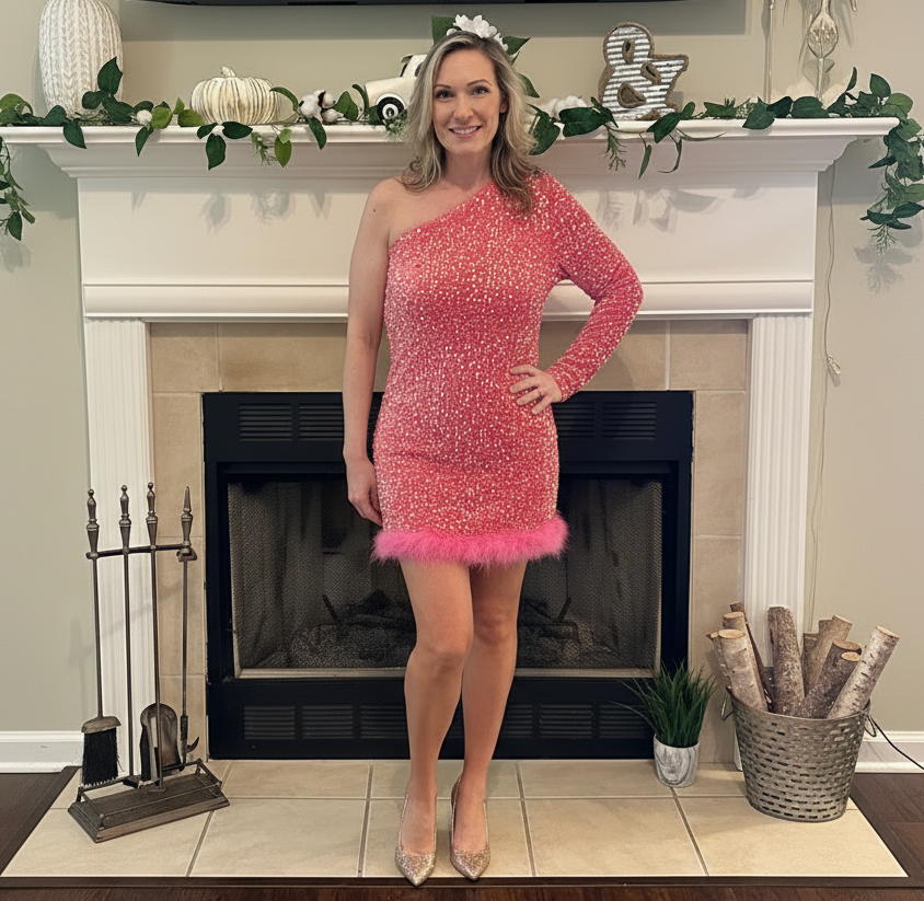 Woman in a pink one-shoulder dress with feather details standing in front of a fireplace.